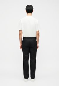 Άτομο με κοντά μαύρα μαλλιά, φορώντας λευκό μπλουζάκι, μαύρο παντελόνι και λευκά αθλητικά παπούτσια, στέκεται με την πλάτη προς το μέρος του θεατή, μπροστά σε ένα απλό λευκό φόντο.