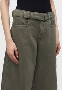 Personne portant un pantalon large taille haute vert olive avec ceinture assortie en tissu et un haut noir, les mains reposant sur les côtés.