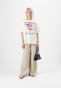 Vit oversized t-shirt med blommig grafik och text, tillsammans med ljusbeiga vida byxor och en liten svart handväska. Bruna slip-on skor.