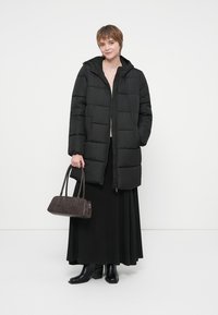 Νεαρή γυναίκα ντυμένη με μαύρο παλτό, μακριά μαύρη φούστα, μαύρες μπότες, κρατώντας μια σκούρα καφέ τσάντα, στέκεται μπροστά σε λευκό φόντο.