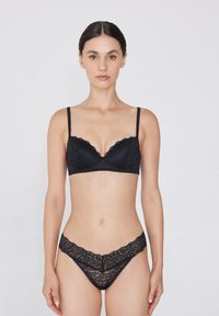 Bralette de encaje negro con bordes con forma de ola, tirantes ajustables y un tanga de encaje a juego. Textura transparente con un patrón floral en toda su superficie.