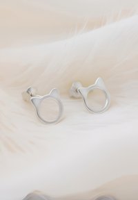 Srebrne kolczyki w kształcie kota z otwartymi okrągłymi detalami i zaokrąglonymi uszami, na miękkim, puszystym białym tle.