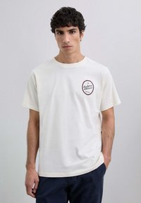 Camiseta corta de manga en color crema, fabricada en tejido suave, con un logo bordado circular en el lado izquierdo del pecho. Cuello redondo, corte relajado y diseño minimalista.