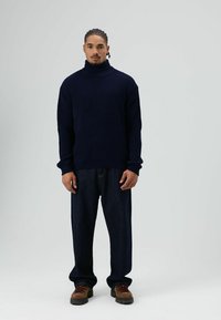 Man met gevlochten haar, gekleed in een marineblauwe coltrui, losse donkere spijkerbroek en bruine wandelschoenen, staand tegen een effen achtergrond.