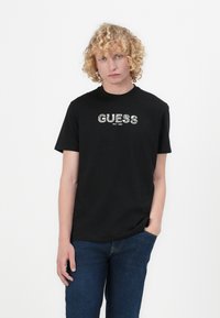 Juodas trumpas rankovėmis marškinėliai iš medvilnės, su tekstūriniu "GUESS EST. 1981" logo baltos spalvos ant krūtinės.