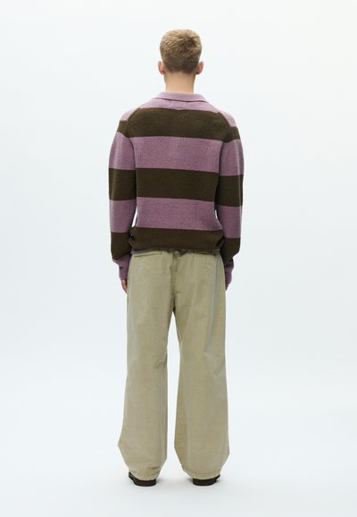 Πλεκτό πουλόβερ με οριζόντιες ριγέ μοβ και καφέ λωρίδες· συνδυάζεται με ανοικτό μπεζ, φαρδιά παντελόνια. Η πίσω όψη αναδεικνύει υφές και σχήματα.