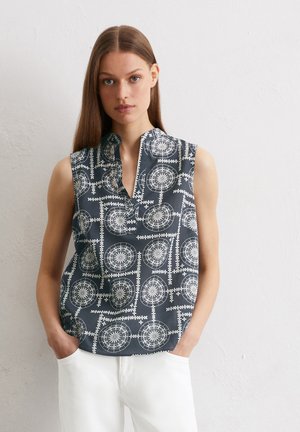 Ärmellose Bluse aus dunklem blauen Stoff mit weißem geometrischen Print und Stehkragen. Das Modell trägt sie mit hellen Hosen.
