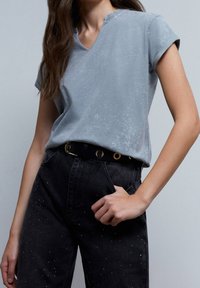 Grå, kortærmet v-hals t-shirt med en blød tekstur, parret med sorte jeans med små hvide sprøjt og et sort bælte med gyldne accenter.