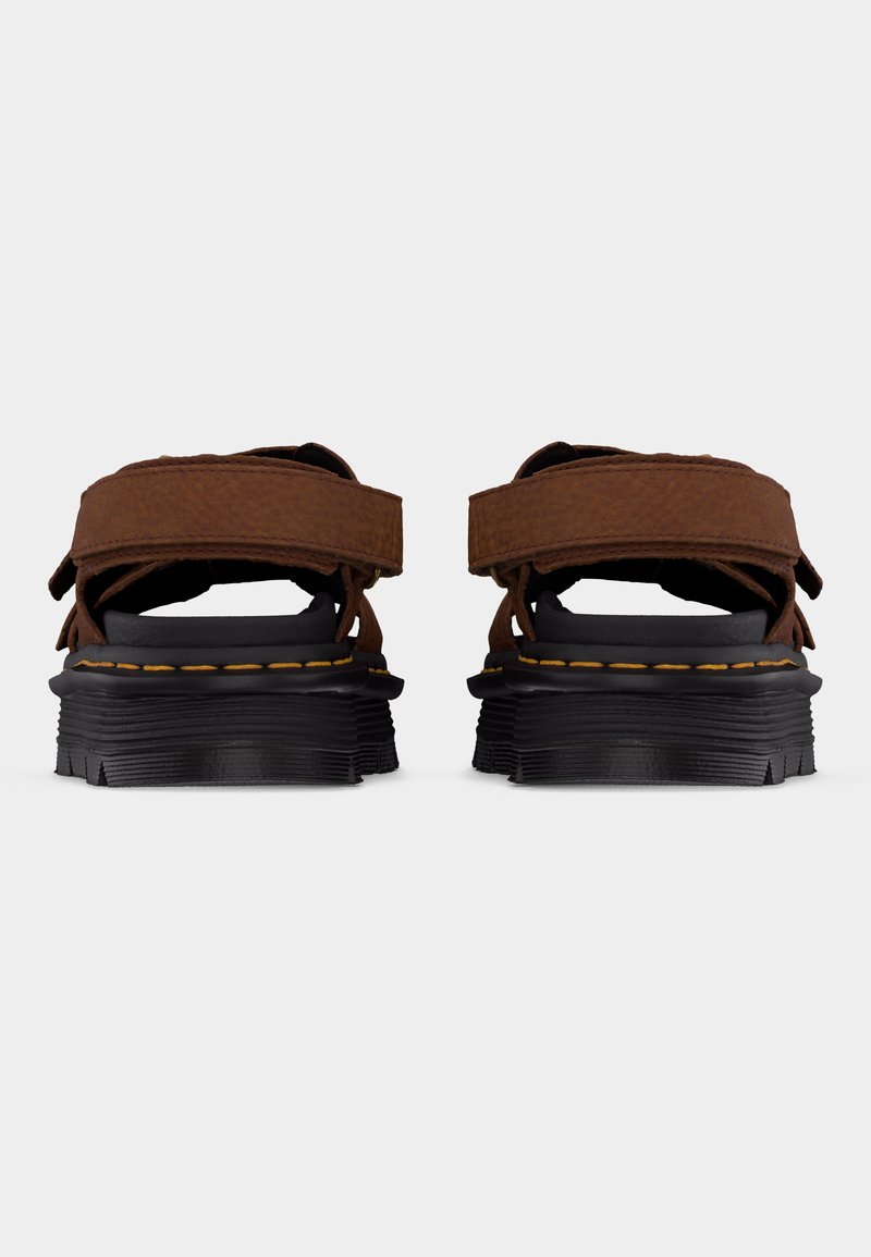 Braune Ledersandalen mit schwarzen, dicken Sohlen und verstellbaren Riemen, von hinten vor einem schlichten hellen Hintergrund gezeigt.