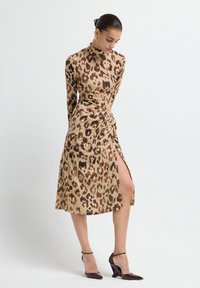 Donna che indossa un vestito a maniche lunghe beige e marrone con stampa leopardata, dotato di uno spacco alto, abbinato a scarpe con tacco alto marroni scuri.