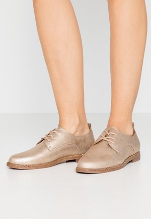 Beige læder oxford-sko med snørebånd, båret på bare fødder mod en ensfarvet lys baggrund.
