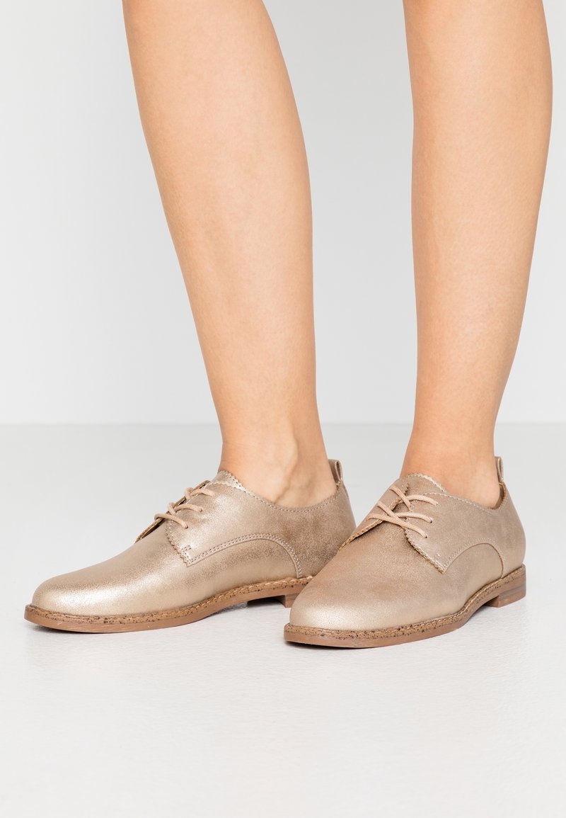 Chaussures Oxford beiges en cuir à lacets portées pieds nus sur un fond clair uni.
