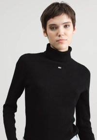 Maglione nero a collo alto a coste con maniche lunghe, presenta un piccolo logo multicolore sul petto. La texture è liscia e aderente.