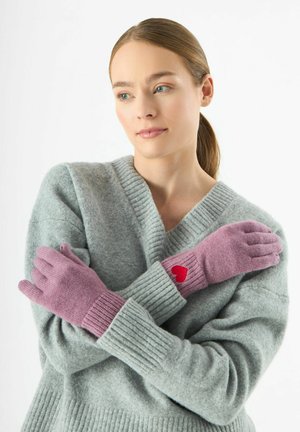 Roze gebreide handschoenen met geribbelde manchetten hebben een klein rood hartdetail. Het model draagt een zachte grijze trui, met gekruiste armen. Eenvoudig, gezellig ontwerp.