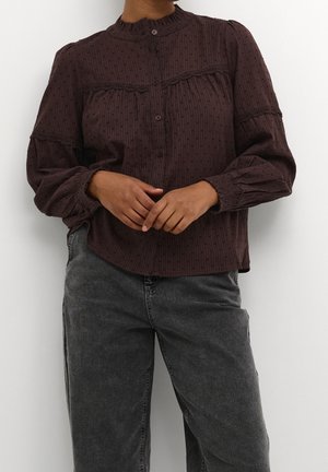 Blusa marrón de botones con tela texturizada, cuello alto y puños de manga elásticos. Combinada con pantalones de mezclilla grises.