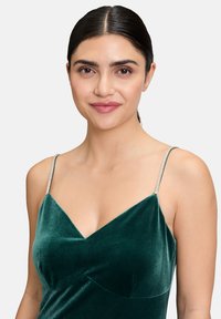 Zielona aksamitna sukienka z dekoltem w kształcie litery V, cienkimi błyszczącymi ramiączkami i miękką fakturą, prezentująca dopasowany krój i subtelne detale.