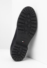 Zwarte rubberen zool met diepe groeven voor grip, geëmbosseerd logo en maatmarkeringen. Het ontwerp heeft een stevige, gestructureerde oppervlakte.
