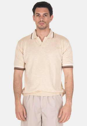 Uomo con capelli corti e scuri che indossa una polo beige a maniche corte con polsini a righe marroni e bianche e pantaloni beige chiaro.