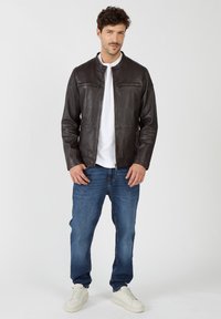 Braune Lederjacke mit Stehkragen, mit Reißverschlüssen versehenen Taschen und tailliertem Design, kombiniert mit blauen Jeans und weißen Sneakers.
