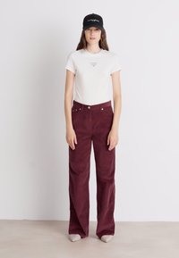 Λευκό κοντομάνικο μπλουζάκι με λογότυπο, μπορντό παντελόνι καμπάνα από χοντρή βελούδινη ύφανση και μαύρο καπέλο. Απλή υφή, χαλαρός σχεδιασμός, ελάχιστες λεπτομέρειες.