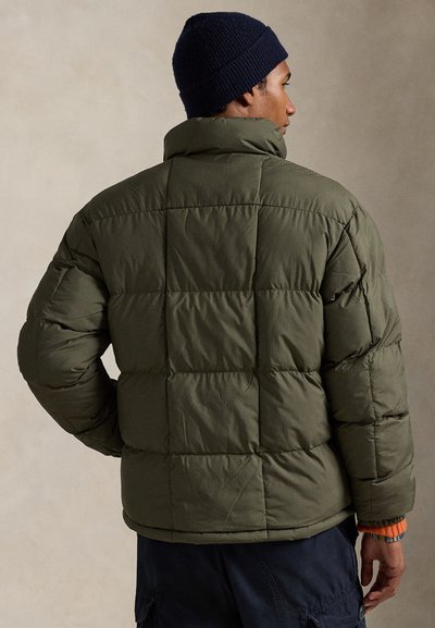 Alyvuogių žalios spalvos pūkinė striukė su apykakle, kvadratinės formos siuvinėjimu ir elastingu apvadu apačioje. Derinama su tamsiai mėlynu megzto audinio kepurėle.