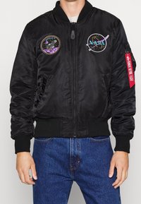 Czarna kurtka bomber z przednim zamkiem, ściągaczami przy mankietach oraz dwoma haftowanymi naszywkami. Posiada boczną kieszeń z czerwoną zawieszką oznaczoną napisem "Remove Before Flight".