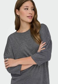 Mujer joven con cabello castaño largo, vistiendo una camiseta gris de manga larga, con los brazos cruzados y mirando de lado con una leve sonrisa.