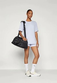 Schwarze Sporttasche mit kurzen Griffen und verstellbarem Tragegurt, ausgestattet mit Reißverschlüssen und einer glatten Textur. Das Modell trägt ein hellblaues T-Shirt und Shorts.