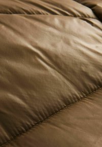 Brun pufferjakke i stof med en blød, glat overflade og subtil glans. Har syede rum, hvilket giver et quiltet udseende.