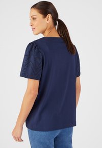 T-shirt bleu marine à manches courtes bouffantes avec des motifs géométriques découpés au laser. Tissu doux et décontracté ; coupe ample. Vue de dos.