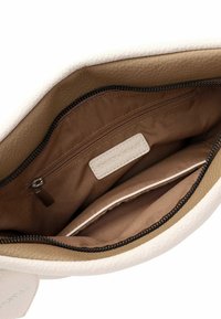 Innenraum einer tanfarbenen und weißen Lederhandtasche mit einem Reißverschlussfach und einem Etikett, das sowohl glatte als auch strukturierte Materialien zeigt. Der Reißverschluss hebt die Funktionalität hervor.