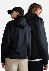 Zwarte nylon jassen met capuchons, voorzien van elastische manchetten en een subtiele logopatch, gedragen door twee modellen die van de camera wegkijken.