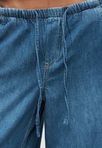 Μπλε τζιν παντελόνι με ελαστική μέση και κορδόνι δέσιμο στο μπροστινό μέρος, φορεμένο σε πρόσωπο με ορατό δέρμα στη μέση από πάνω.