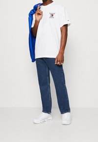 Camiseta blanca de algodón con el logo rojo de "Tommy Jeans" en el lado izquierdo del pecho, combinada con jeans de mezclilla azul oscuro y zapatillas blancas, con una chaqueta azul.