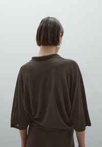Brown, draped tunic top with a shawl collar and loose sleeves, featuring a smooth texture and a relaxed fit. Background is neutral gray.