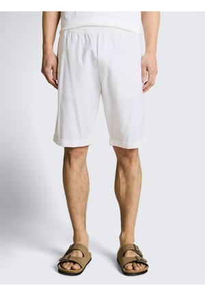 Man draagt wit knielang korte broek en bruine sandalen, staand op een effen lichte achtergrond.