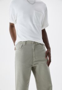 Homme portant un t-shirt blanc à manches courtes avec une poche sur la poitrine et un pantalon gris clair, les mains détendues le long du corps, avec un collier chaîne argenté.