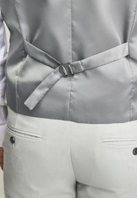 Gilet gris clair avec une finition satinée lisse, doté d'une boucle arrière et d'une sangle en satin. Associé à un pantalon gris clair texturé avec poches.