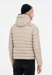 Doudoune beige avec un matelassage horizontal, une capuche ajustée et un logo noir sur l'épaule. Portée avec un jean foncé et un bonnet.