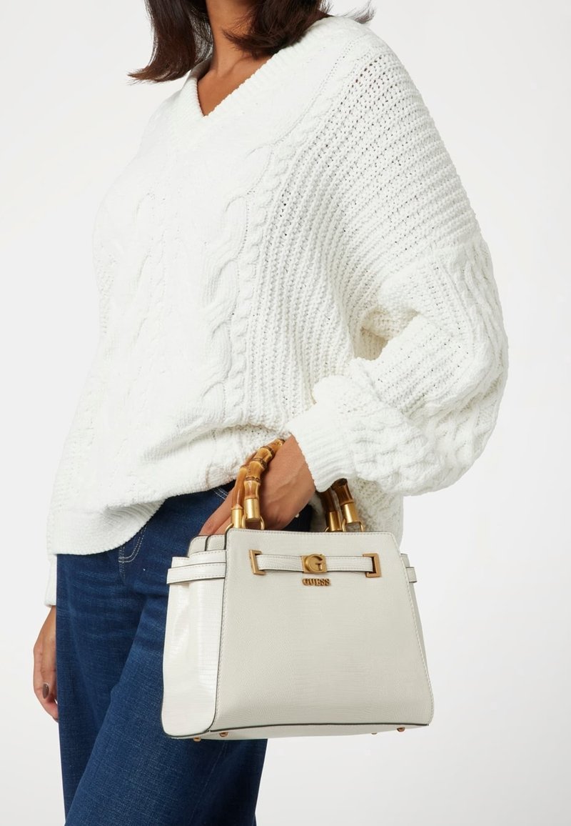 Biała torebka na ramię z bambusowymi uchwytami, złotymi detalami i teksturowaną powierzchnią, noszona obok osoby w białym swetrze i niebieskich dżinsach.