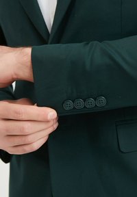 Blazer vert foncé avec une texture lisse, comportant quatre boutons noirs sur le poignet. La manche est légèrement retroussée pour révéler des détails.