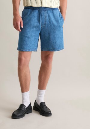 Lichtblauwe denimshorts met een elastische tailleband en zijzakken, gecombineerd met zwarte instapschoenen en witte crew-sokken.