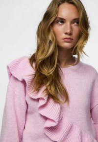 Różowy sweter na drutach z wyraźnym falbaną na lewym ramieniu, o luźnym kroju i miękkiej teksturze.