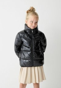 Giacca nera imbottita con colletto alto e texture lucida, dotata di una zip frontale e dettagli di cucitura orizzontale, indossata sopra una gonna beige.
