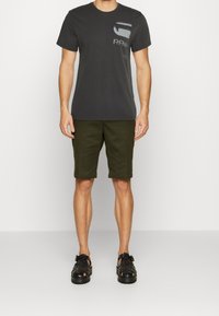 Šedé tričko s designem kapsy, olivově zelené šortky a černé sandály. Hladký materiál s volným střihem. Obsahuje akcenty loga.