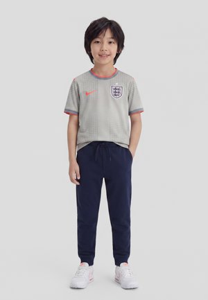 Niño pequeño de pie con camiseta de fútbol gris de Inglaterra, pantalones deportivos azul marino y zapatillas blancas con detalles en rosa sobre fondo liso.