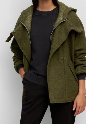 Persona con sudadera gris oscuro bajo una chaqueta de lana verde oliva con cremallera, mangas remangadas y pantalones negros, de pie con una mano en el bolsillo.