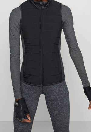 Personne portant une veste matelassée noire sans manches sur un haut gris à manches longues et un pantalon gris ajusté, avec des gants noirs sans doigts.