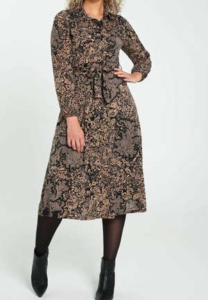 Kobieta ubrana w czarną i beżową sukienkę midi w kwiatowy wzór z długim rękawem i paskiem do wiązania, czarne rajstopy i czarne botki.