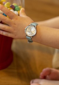 Zilveren horloge met grote cijfers op een witte wijzerplaat, lichtblauwe lederen band, gedragen om de pols van een kind, met een kleurrijke snoepjespot op de achtergrond.